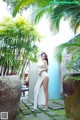 A woman in a white dress standing in front of a palm tree.